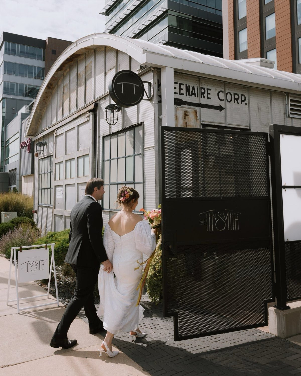 Bride and groom walking toward an eclectic industrial venue in a city setting.
