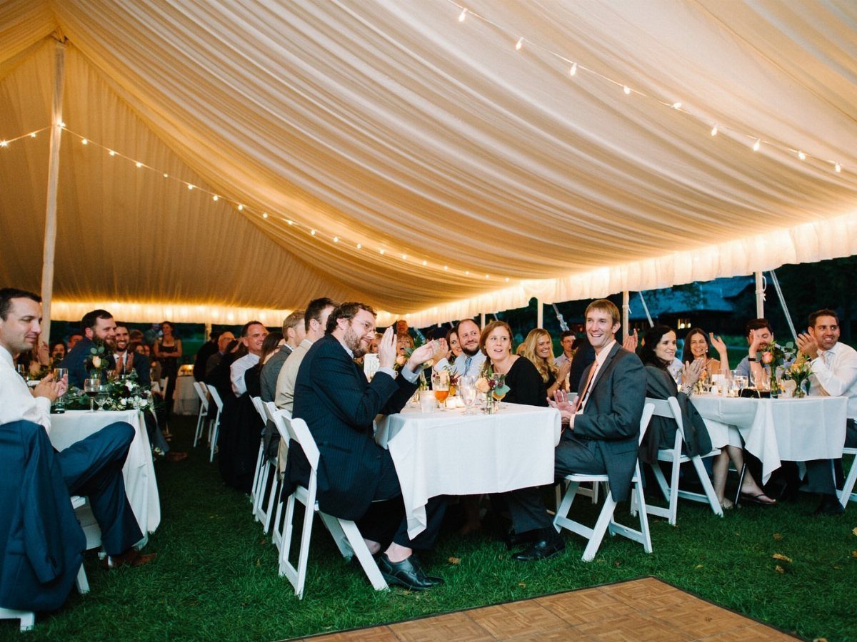 Wedding guests dining at tables under a large white tent with string lights.
