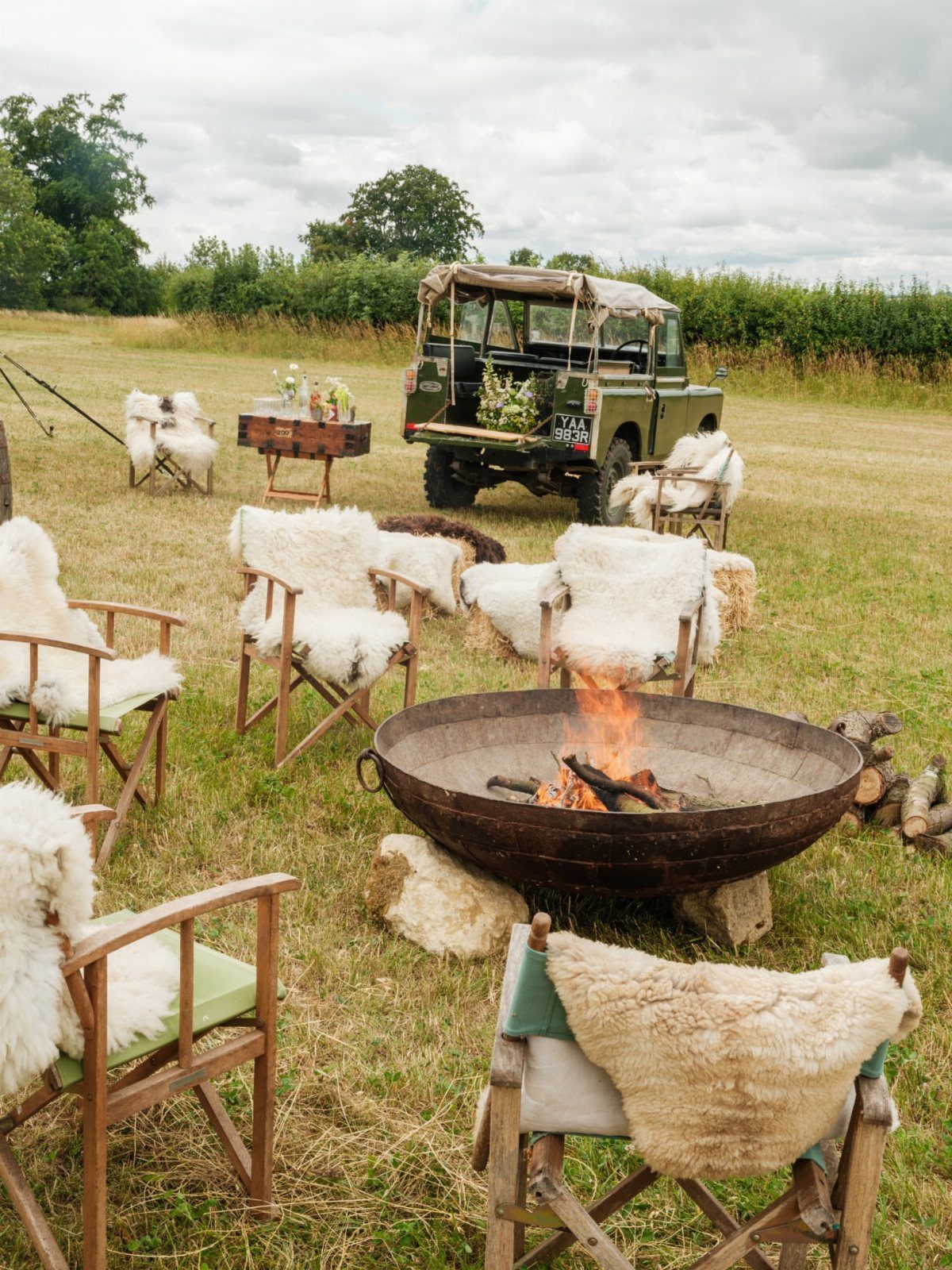 Outdoor lounge with fire pit, sheepskin chairs, and vintage jeep in a field