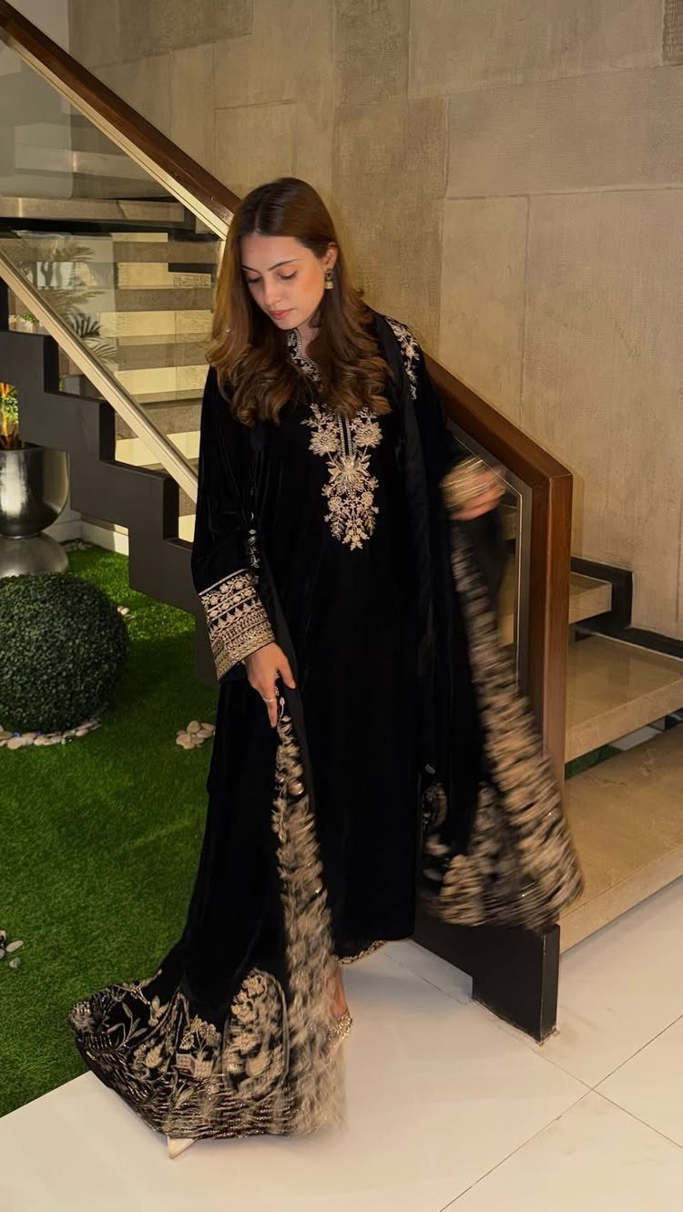 Woman in a black velvet dress with gold embroidery standing by a staircase.