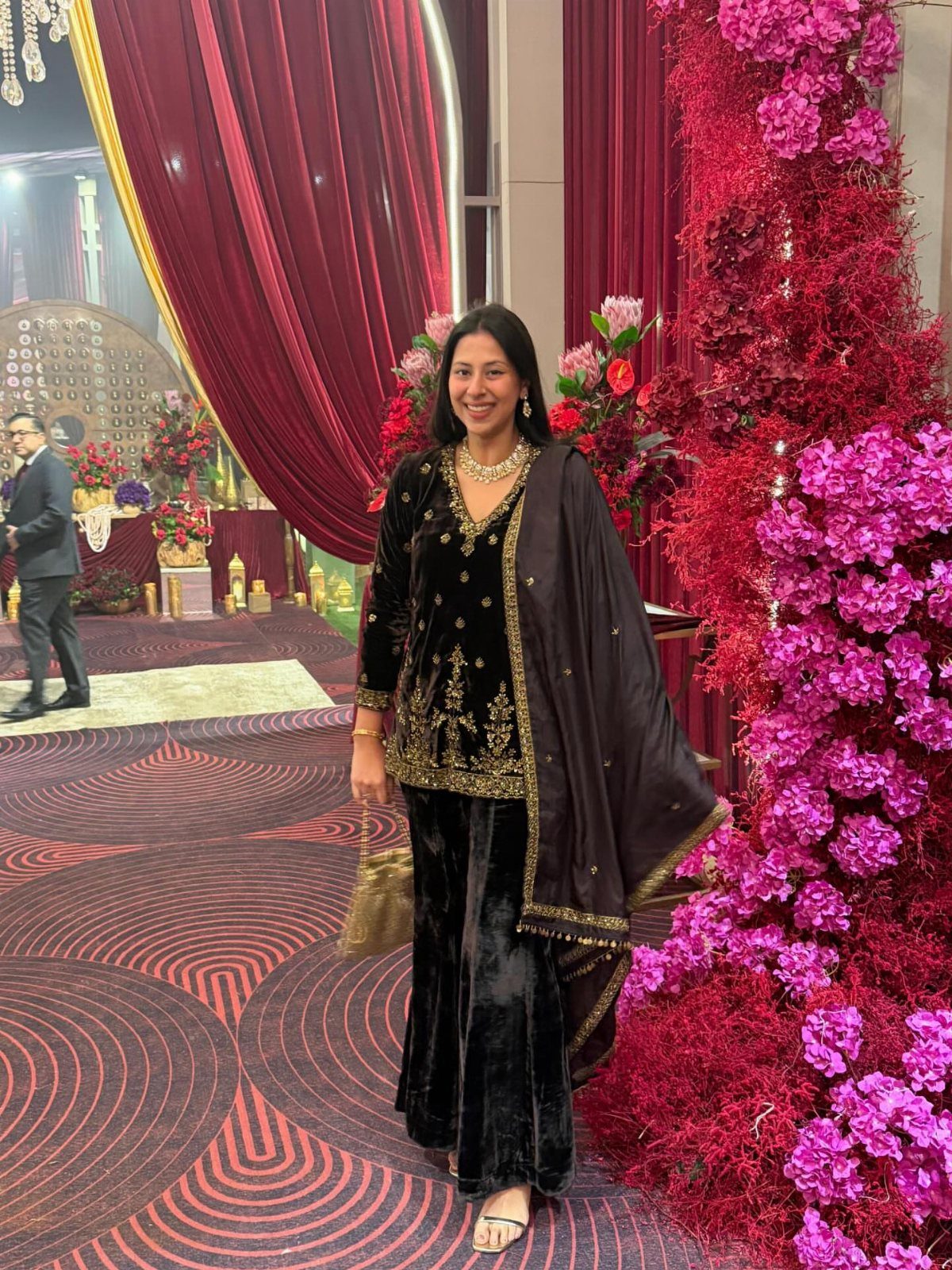 Woman in black velvet embroidered traditional Indian outfit standing by pink floral decorations.